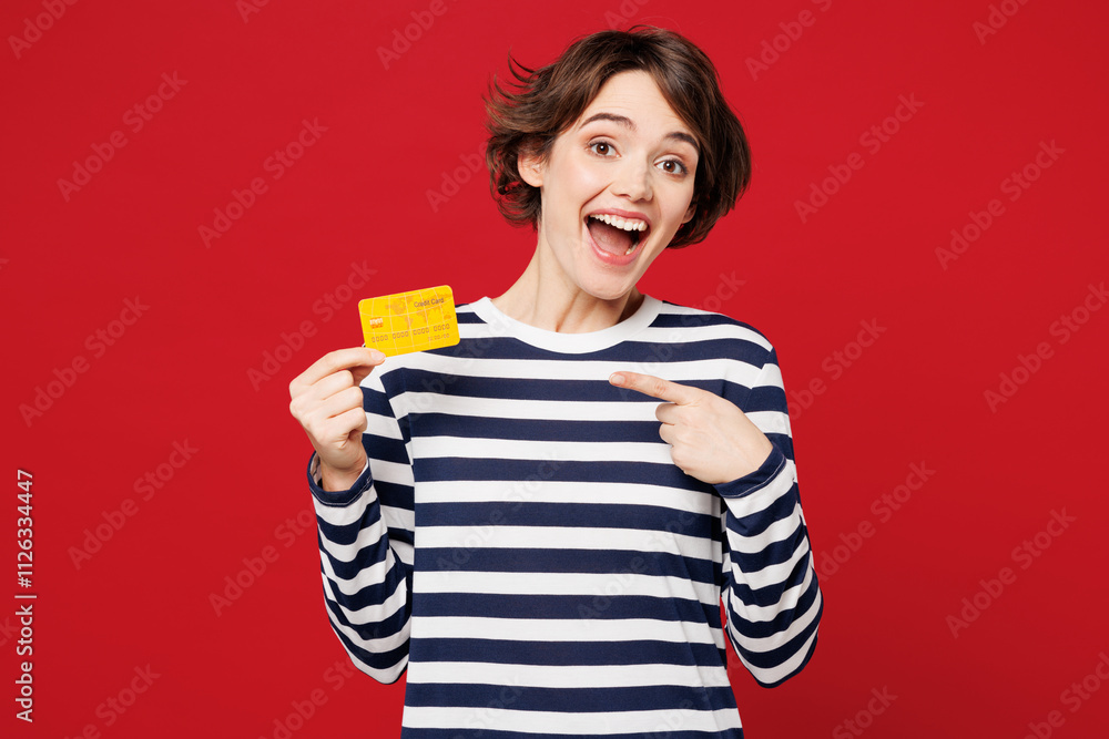 Young surprised shocked woman wear blue striped sweatshirt casual clothes hold in hand point finger on mock up of credit bank card isolated on plain red background studio portrait. Lifestyle concept. Young surprised shocked woman wear blue striped sweatshirt casual clothes hold in hand point finger on mock up of credit bank card isolated on plain red background studio portrait. Lifestyle concept.