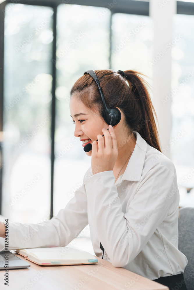 smiling woman wearing headset works at desk, engaging in professional conversation. She is dressed in white shirt, with notepad and laptop in front of her, in bright office smiling woman wearing headset works at desk, engaging in professional conversation. She is dressed in white shirt, with notepad and laptop in front of her, in bright office