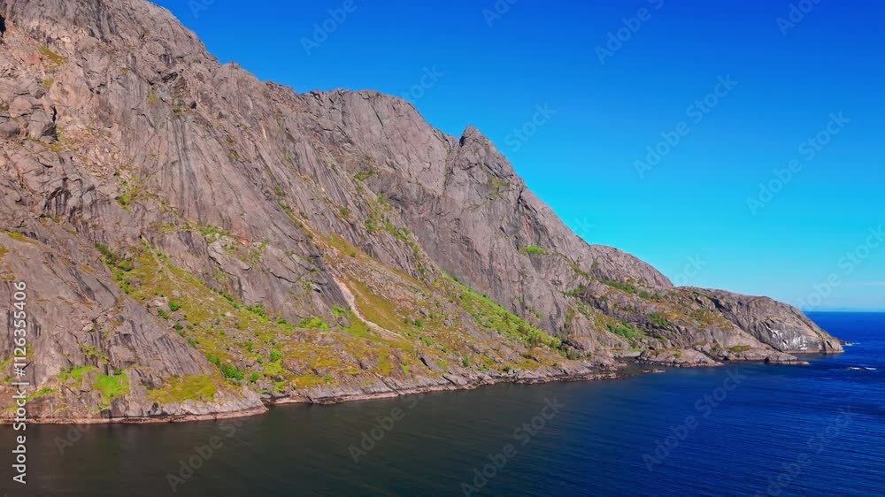 A large fjord side mountain rock face in Nusfjord, Lofoten, northern Norway. Green vegetation on sides of cliffs, open sea in the background. Aerial shot approaches, sunny with blue sky. A large fjord side mountain rock face in Nusfjord, Lofoten, northern Norway. Green vegetation on sides of cliffs, open sea in the background. Aerial shot approaches, sunny with blue sky.