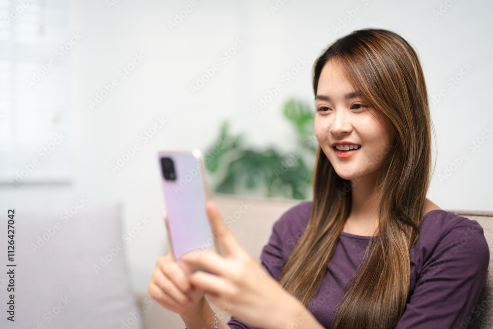 Asian woman sit on sofa holding phone looking at cellphone screen waving hand video calling distance friend online in mobile chat app using smartphone video chat application. distant web camera call. Asian woman sit on sofa holding phone looking at cellphone screen waving hand video calling distance friend online in mobile chat app using smartphone video chat application. distant web camera call.