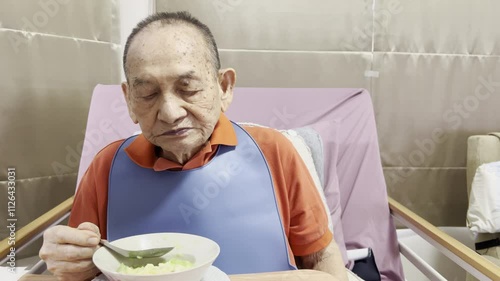 An elderly man seated in a care facility, wearing a protective bib and eating from a bowl. The image conveys themes of elderly care, health, and support in a nurturing environment