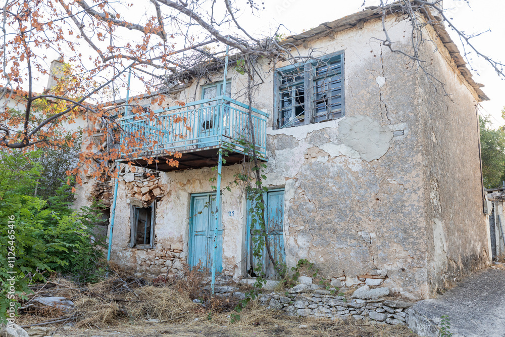 An old, abandoned house with faded blue shutters and a crumbling facade, surrounded by wild greenery in a traditional Greek village. An old, abandoned house with faded blue shutters and a crumbling facade, surrounded by wild greenery in a traditional Greek village.