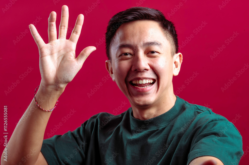 Asian man in a green T-shirt smiling while holding a smartphone in one hand, raised for a selfie or video call, and waving with the other hand, against a vibrant red background Asian man in a green T-shirt smiling while holding a smartphone in one hand, raised for a selfie or video call, and waving with the other hand, against a vibrant red background