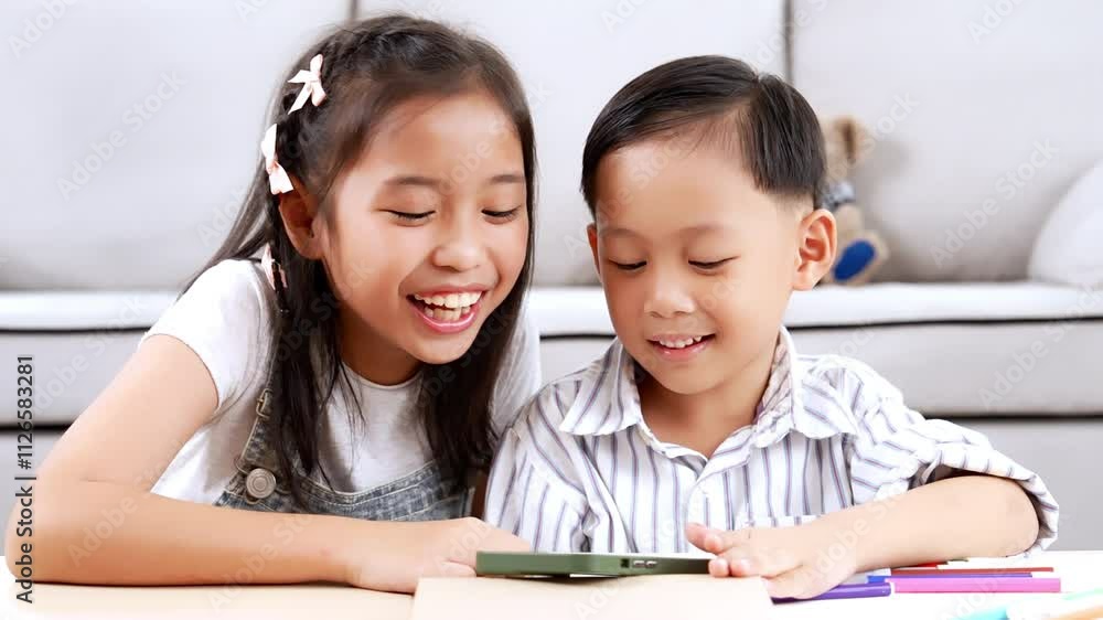 Asian elder sister and younger brother using mobile phones to watch movies or cartoons in living room. They enjoying and having fun with entertainment on phones, using home internet Asian elder sister and younger brother using mobile phones to watch movies or cartoons in living room. They enjoying and having fun with entertainment on phones, using home internet
