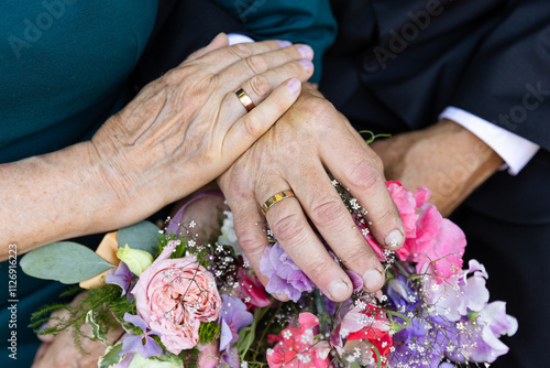 Two hands, worn with age, hold each other gently, showing their wedding rings that have lasted through the years. The rings and flowers tell a story of a lifelong journey together, full of love