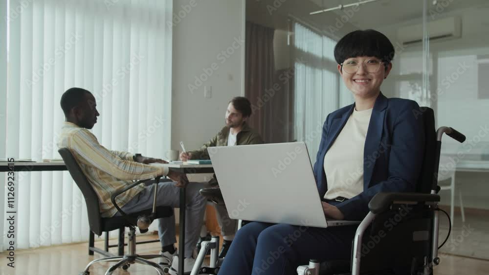 Low angle portrait of Asian female manager in wheelchair smiling at camera while working on laptop in inclusive coworking space Low angle portrait of Asian female manager in wheelchair smiling at camera while working on laptop in inclusive coworking space