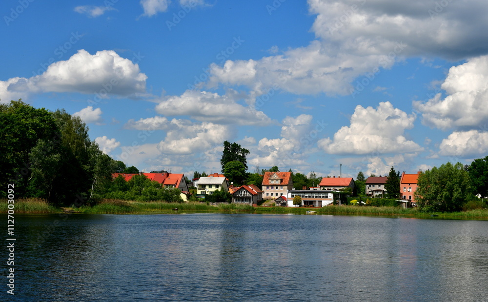 Obraz premium A view of a vast yet shallow river, lake or channel surrounded from all sides with reeds, shrubs and forests with some stone and concrete barriers seen, a small jetty, some boats and a quaint town