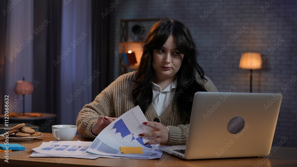 Young business woman freelancer sitting at desk working on remote in home office late, looks at laptop, holding searching through documents. Young business woman freelancer sitting at desk working on remote in home office late, looks at laptop, holding searching through documents.