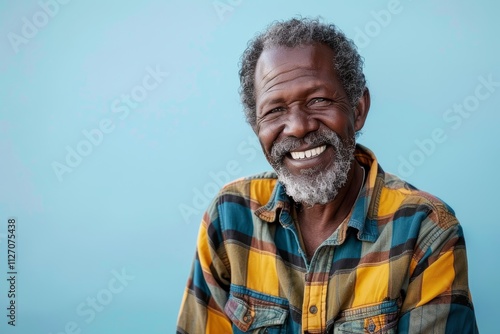 Portrait of a grinning afro-american man in his 70s dressed in a relaxed flannel shirt in front of soft blue background