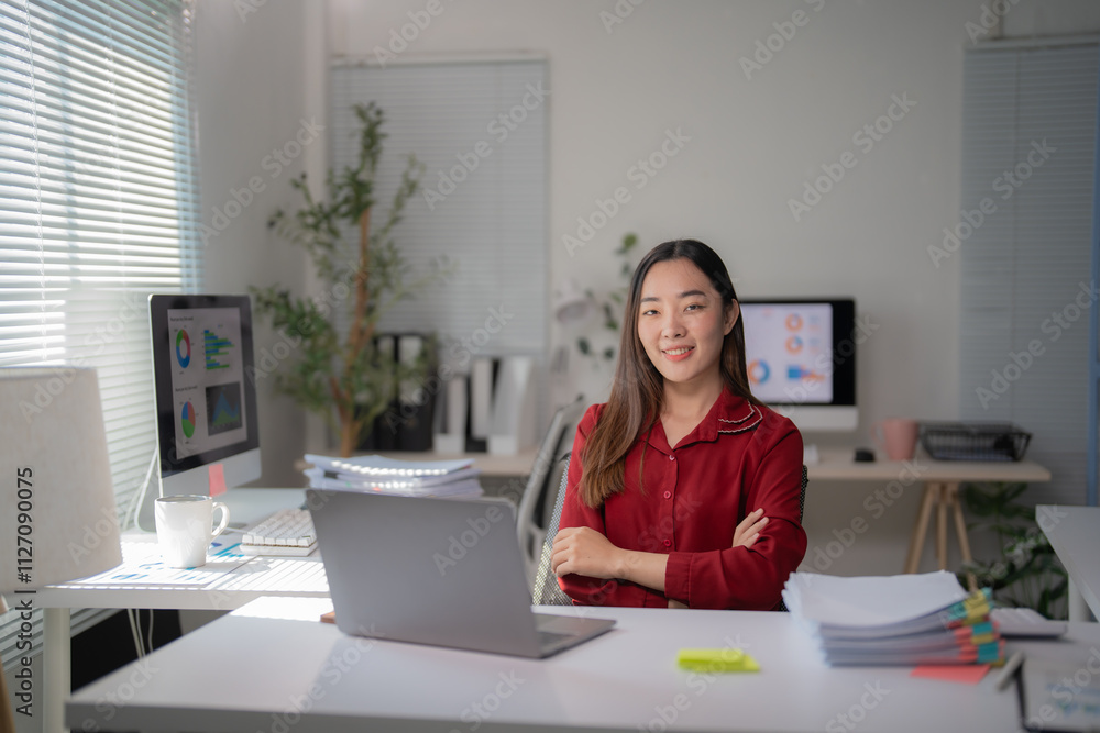 Confident young asian businesswoman smiling with arms crossed sitting at office desk working using laptop computer with documents and computer showing graphs Confident young asian businesswoman smiling with arms crossed sitting at office desk working using laptop computer with documents and computer showing graphs