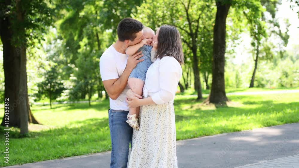 Cute family going for walk in park, mother and father carrying baby in arms. Parents kissing toddler. Happy family moments, summer walk outside. International Family Day. Cute family going for walk in park, mother and father carrying baby in arms. Parents kissing toddler. Happy family moments, summer walk outside. International Family Day.