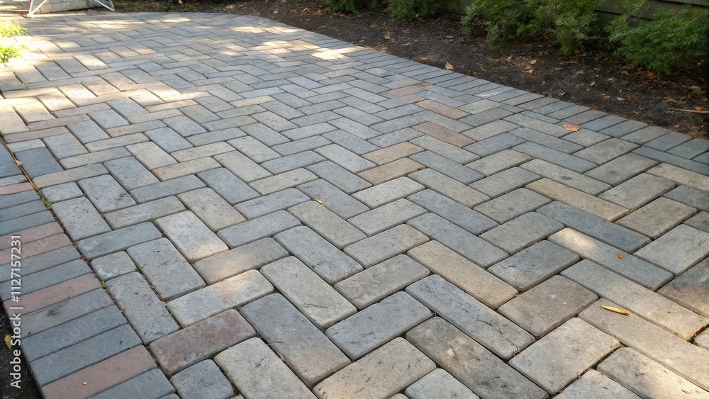 Fototapeta premium A herringbone pattern of gray, tan, and red bricks creates a walkway lined with lush greenery.