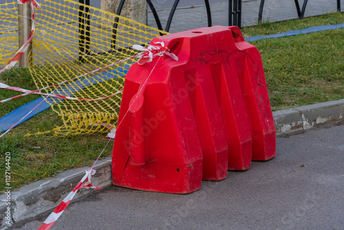 A vivid red plastic barrier stands prominently on the roadside, cordoned off with caution tape as workers prepare for upcoming construction. Sunlight illuminates the area, enhancing visibility photo