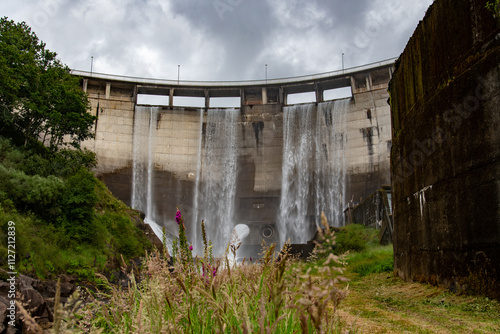 Wallpaper Mural The image shows a large concrete dam with water cascading down its face from multiple spillways. The structure is massive and surrounded by lush green vegetation and cloudy skies. Torontodigital.ca