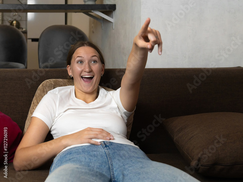 Close up young woman lying on a comfortable sofa, pointing her finger at the tv screen with a cheerful expression, enjoying a relaxing moment of home entertainment