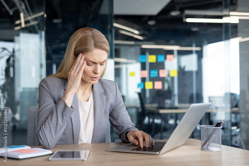 A businesswoman experiencing stress and headache works on her laptop in a modern office setting, surrounded by technology and colorful notes. A businesswoman experiencing stress and headache works on her laptop in a modern office setting, surrounded by technology and colorful notes.