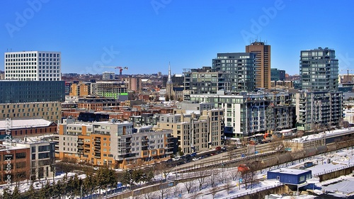 A vibrant cityscape showcasing modern architecture and urban development under a clear blue sky. Snow-covered streets and rooftops add a seasonal charm to the bustling metropolitan area.