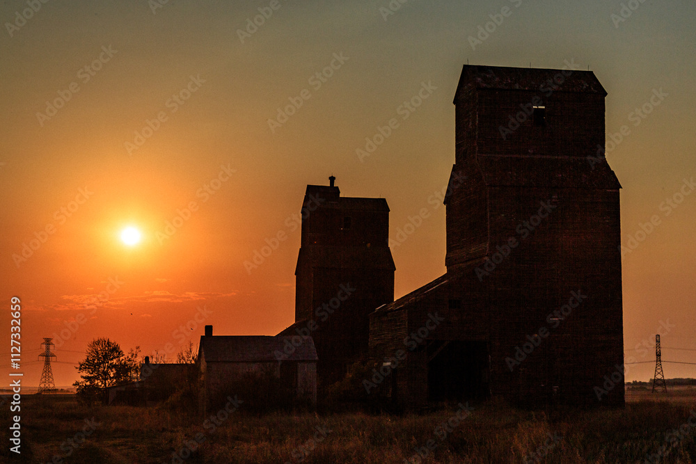 Obraz premium The sun is setting over a field with two old grain silos