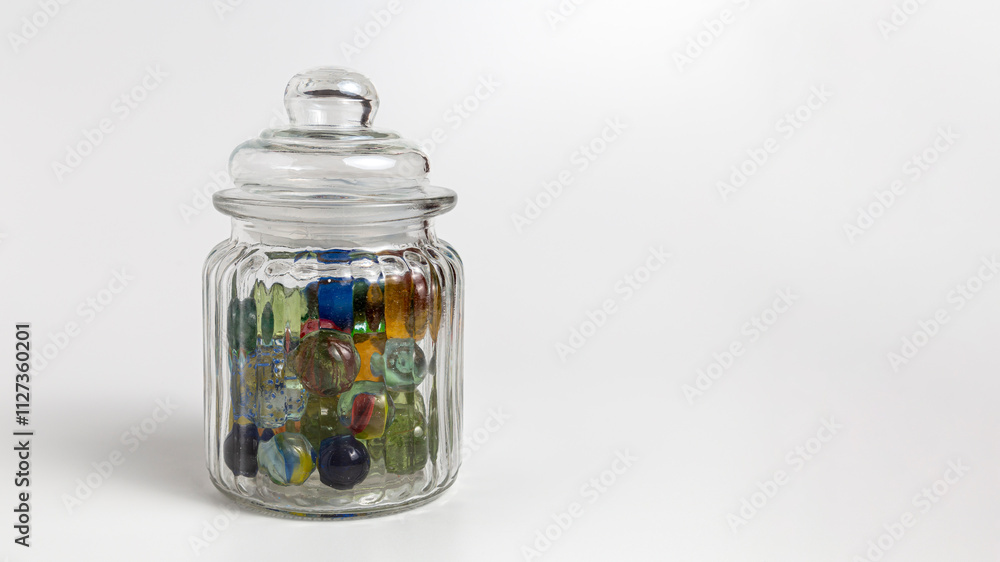 Multicolored glass marbles in a glass jar with lid on a white background. Copy space.
Multicolored glass marbles in a glass jar with lid on a white background. Copy space.