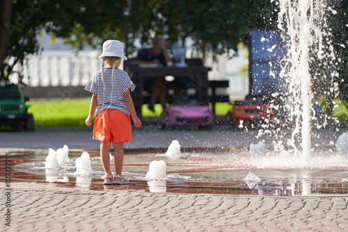 Cute little girl in sailor striped T-shirt on a hot day at a fountain with splashing and splashing water. In the background blurred elements of the park infrastructure. photo