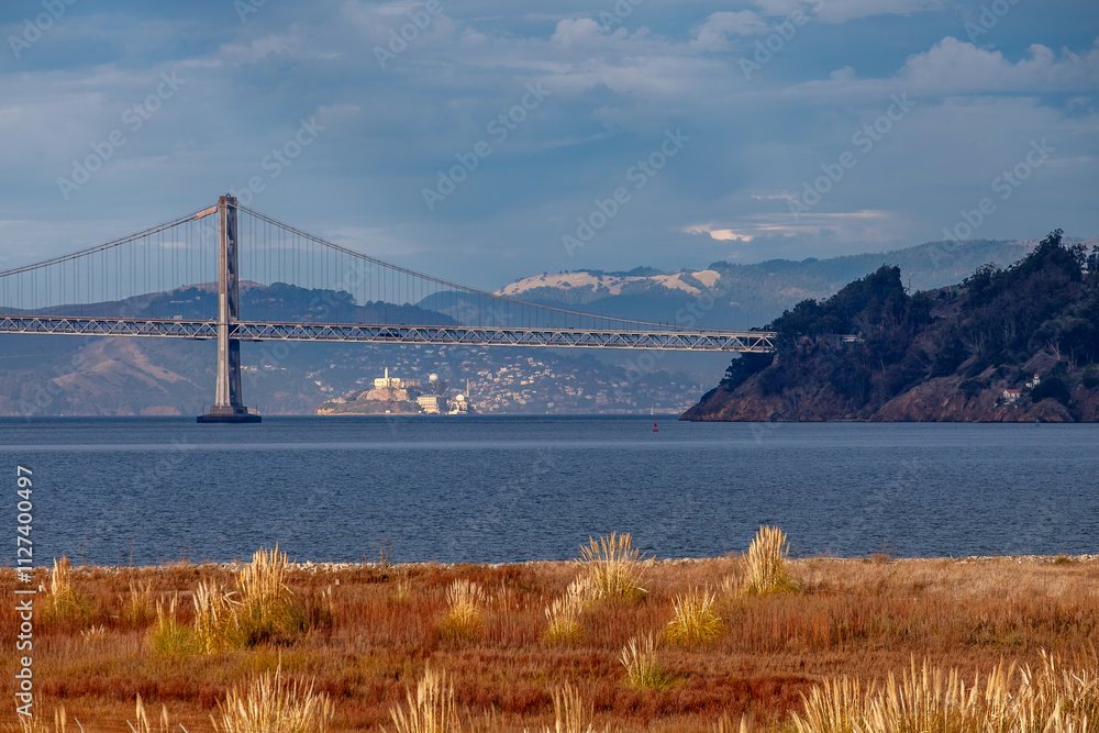 Naklejka premium The photograph captures the serene morning atmosphere with calm waters of the San Francisco Bay, the iconic bridge structure, and the historic Alcatraz Island.