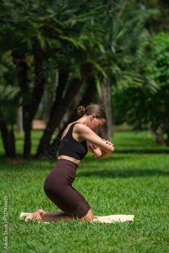 Slender young woman in sports clothes kneads her body on a fitness mat in a public park, does yoga. Woman takes care of her mental and physical health. Vertical photo