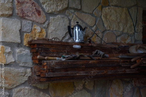 A metal kettle on a rustic wooden shelf with tools, set against a textured stone wall, evoking a countryside or workshop setting