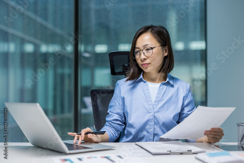 Asian woman businesswoman engaged in work at her desk, using a laptop and reviewing documents. The office setting highlights productivity and professionalism, reflecting a focused and organized work photo