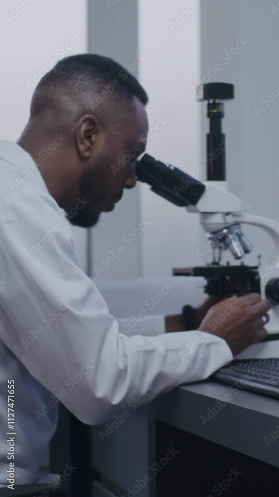Vertical shot African American microbiologist looks under microscope, does analysis of test sample using high-tech equipment. PC screen shows image of cells or bacteria. Microbiology. Vertical shot African American microbiologist looks under microscope, does analysis of test sample using high-tech equipment. PC screen shows image of cells or bacteria. Microbiology.