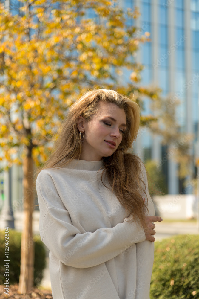 Portrait of beautiful smiling serene long haired blond caucasian woman in white sweater standing outdoors on city street in a sunny autumn day by tree with yellow leaves. Soft focus. People theme. Portrait of beautiful smiling serene long haired blond caucasian woman in white sweater standing outdoors on city street in a sunny autumn day by tree with yellow leaves. Soft focus. People theme.