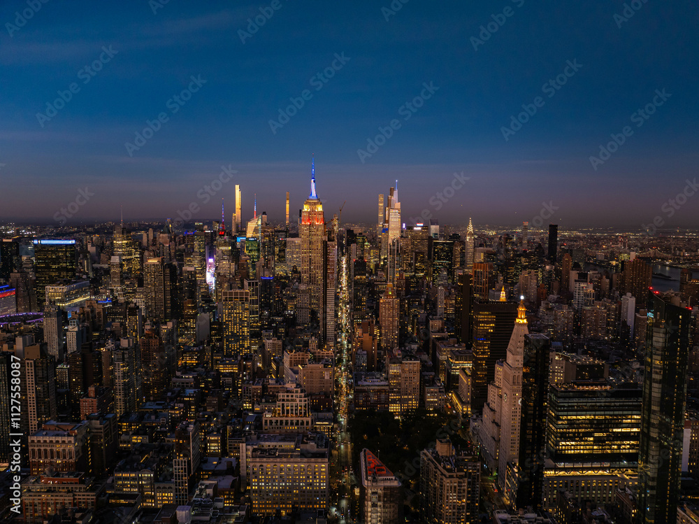 Fototapeta premium Illuminated New York City skyline at twilight, featuring the iconic Empire State Building amid a vibrant urban landscape of towering skyscrapers in Midtown Manhattan