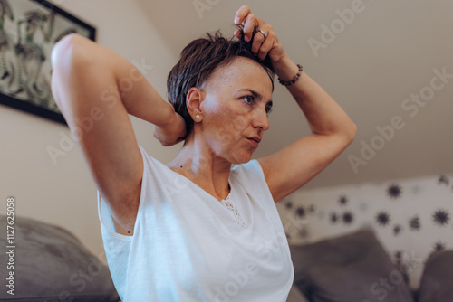 A woman with cancer sits on a couch, adjusting her wig with confidence, symbolizing self-care and connection to her personal style in a cozy environment. photo