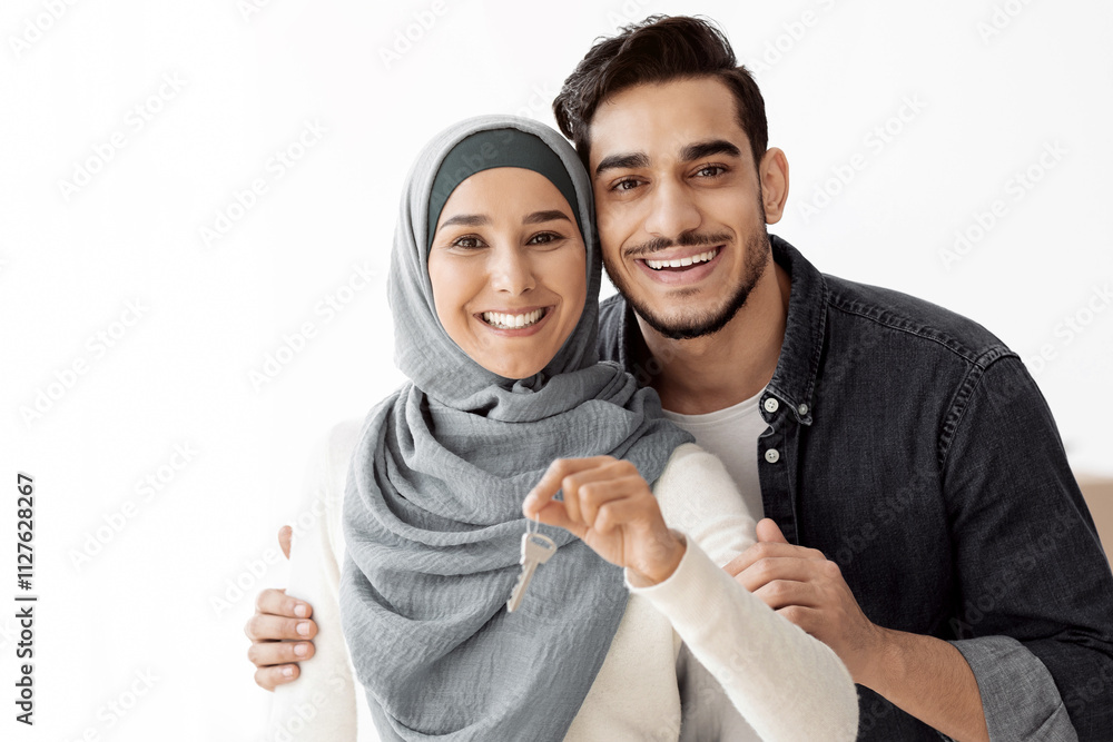 Happy young arab family husband and wife embracing and showing apartment key, copy space. Handsome middle-eastern man hugging his beautiful woman in hijab, moving to new house, relocation concept Happy young arab family husband and wife embracing and showing apartment key, copy space. Handsome middle-eastern man hugging his beautiful woman in hijab, moving to new house, relocation concept