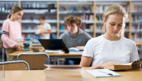 Portrait of a fifteen-year-old schoolgirl sitting at a desk in the school library, attentive reading a book