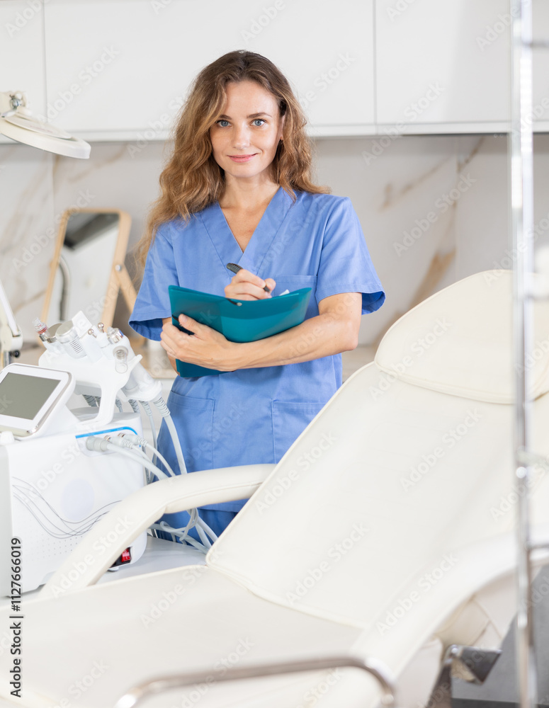 Fototapeta premium Young female cosmetologist in medical uniform taking notes in folder in medical room
