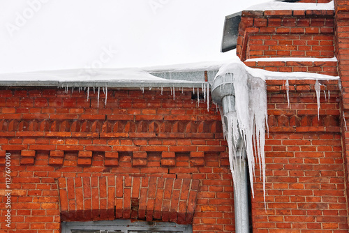 Icicles on gutter and roof. Huge Icicles hanging down from drain pipe of the red building. Icy facade of building, dangerous icicles melt on rooftop, risk of injury. Ice hazard