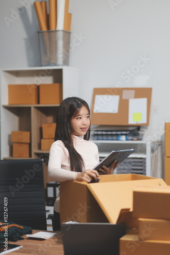 Startup SME small business entrepreneur of freelance Asian woman wearing apron using laptop and box to receive and review orders online to prepare to pack sell to customers, online sme business ideas.