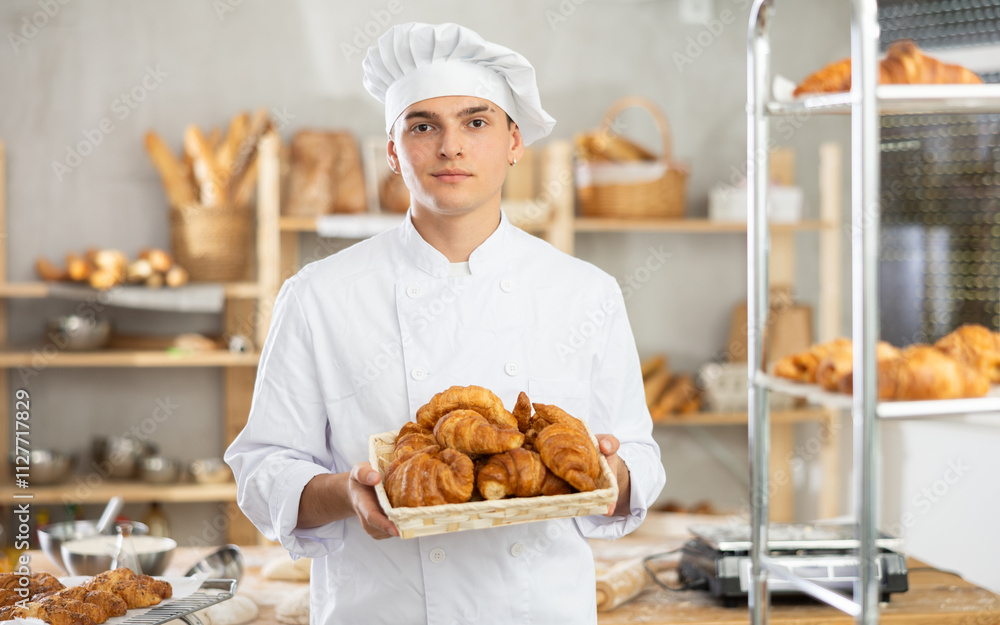 Positive young male baker holding croissants in square wicker basket in bakery shop Positive young male baker holding croissants in square wicker basket in bakery shop