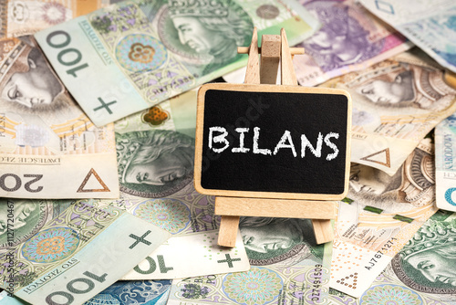 Black writing board on a wooden frame with the inscription "Bilans", Polish zloty PLN banknotes scattered in the background (selective focus) translation: Balance