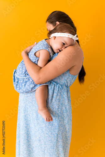 Motherhood, hot summer - portrait young pregnant happy smiling joy mother woman in blue dress holding little baby daughter toddler sibling bare feet in arms looking into eyes kiss on yellow background