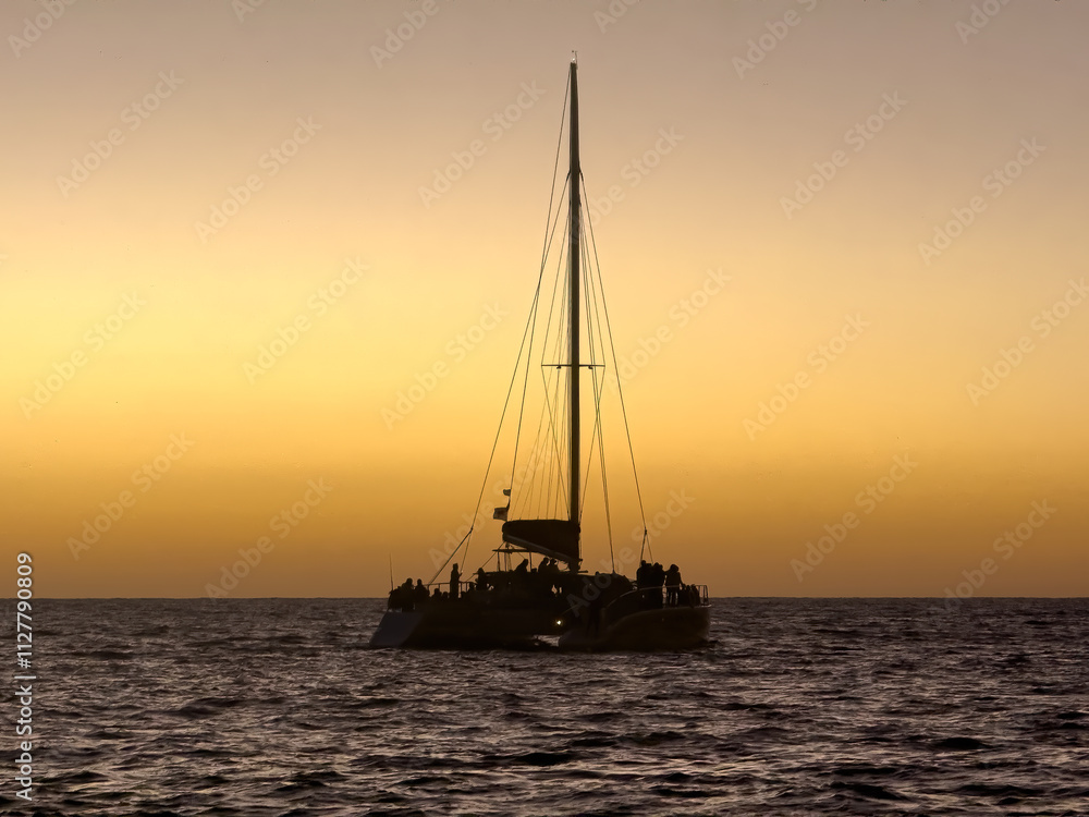 A catamaran, silhouetted against a fiery sunset, glides across the calm waters. The boat is filled with passengers enjoying the breathtaking view, creating a serene and picturesque scene. A catamaran, silhouetted against a fiery sunset, glides across the calm waters. The boat is filled with passengers enjoying the breathtaking view, creating a serene and picturesque scene.