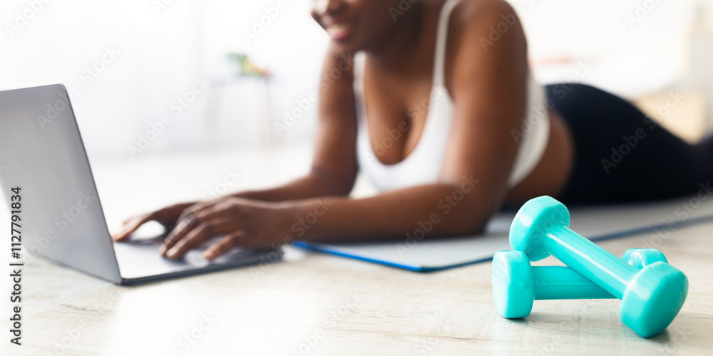 Fototapeta premium Plump young black woman lying on yoga mat and watching online sports video on laptop at home, focus on dumbbells. Plus size millennial lady taking break from domestic training, panorama with copyspace