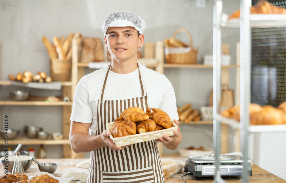 Friendly young male baker carrying square basket with fresh croissants in bakery Friendly young male baker carrying square basket with fresh croissants in bakery