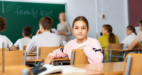 Young girl turned around and looking at camera while sitting at desk during lesson in classroom.
