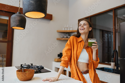 Bild auf Leinwand Young woman enjoying a healthy green smoothie in a bright kitchen, wearing an or