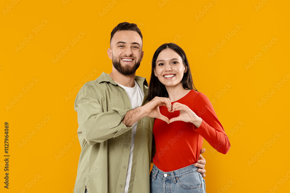 Obraz premium A young couple stands together against a vibrant yellow backdrop, smiling and making a heart shape with their hands. Their affectionate pose reflects their love and happiness.