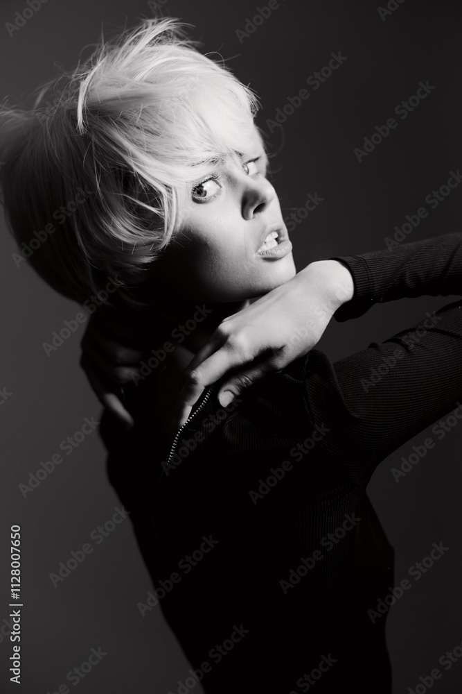 Expressive young woman with short blonde hair, posing dramatically against a dark background, showcasing strong emotions and fashion sense. Artistic black and white portrait. Expressive young woman with short blonde hair, posing dramatically against a dark background, showcasing strong emotions and fashion sense. Artistic black and white portrait.