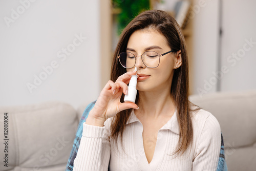 Young woman wearing casual clothing, using a nasal spray to facilitate breathing, sitting on a sofa in a home interior