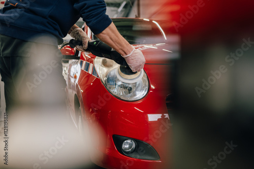 Ein Autopflege-Spezialist poliert den Scheinwerfer eines roten Autos mit einer professionellen Poliermaschine. Die Szene zeigt die Präzision und Detailarbeit bei der Fahrzeugaufbereitung.