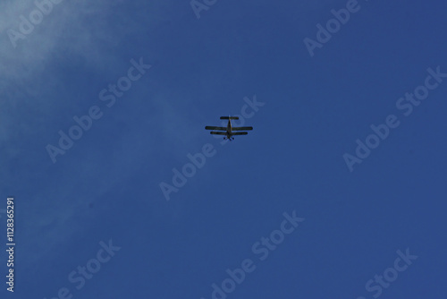 A small motor plane with two wings flies away into the distance in the middle of the blue sky.